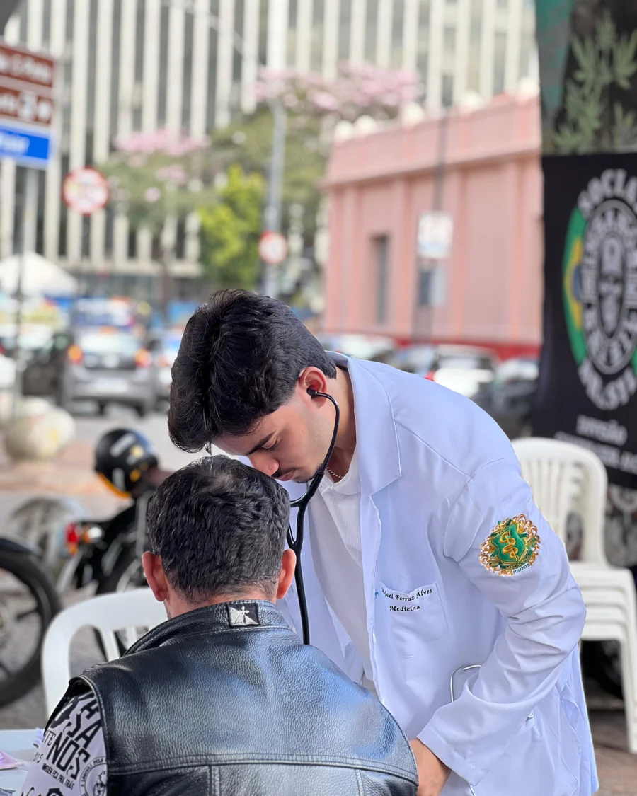 Rafael Ferraz, de jaleco, auscultando um paciente numa ação de saúde na rua